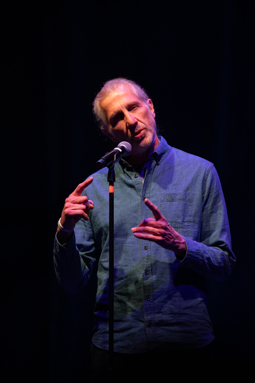 An expressive medium shot of a man speaking or performing on a dimly lit stage.Subject and PosePerson: A middle-aged to older man with light skin, graying hair pulled back, and a short, trim gray beard and mustache.Expression & Pose: He is tilting his head slightly to his left with a focused, expressive facial expression, as if speaking, telling a story, or singing.Gestures: His hands are raised in active gestures. His right hand is raised to about chest height with his index finger pointing upward, while his left hand is held slightly lower, fingers partially extended as if emphasizing a point.Clothing and EquipmentAttire: He is wearing a long-sleeved, button-down blue chambray or denim shirt over a dark undershirt. He has a thin white or light-colored band around his right wrist.Microphone: A microphone on a tall black stand is positioned directly in front of him, close to his mouth. A small band of orange tape is visible near the top of the microphone stand.Lighting and BackgroundLighting: The scene is lit with dramatic stage lighting. A strong spotlight catches the right side of his face and shirt, while the rest of his body is bathed in a cool, deep blue and purple ambient hue.Background: The background is entirely dark and minimalist, keeping the full focus on the performer.Photo credit: kmr studios