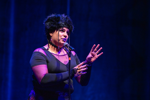 An expressive medium shot of a person speaking or performing live on stage.Subject and PosePerson: A person with light-to-medium skin, distinctive purple lipstick, and dark nail polish. Their dark hair features several thin, beaded braids framing the face.Expression & Pose: They are positioned slightly to the left of the frame, looking and gesturing towards the right with an earnest, focused expression as they speak into the microphone.Gestures: Both hands are raised in front of them to chest-and-shoulder height. Their fingers are spread wide in an open, fluid gesture, adding an dynamic, storytelling quality to the moment.Clothing and AccessoriesAttire: They are wearing an edgy, dark ensemble featuring a black cold-shoulder top over a sheer mesh neckline, paired with long black arm warmers that cover the forearms. A hint of a purple plaid or checkered skirt is visible at the waist.Accessories: They wear a large, prominent black furry hat, a black spiky choker necklace around their neck, and dangling earrings.Equipment: A microphone on a tall black stand is positioned directly in front of them.Lighting and BackgroundLighting: The stage is illuminated by dramatic theatrical lighting. A warm spotlight hits the speaker from the front, highlighting their face and hands, while the rest of the scene is bathed in deep blue and purple tones.Background: The background consists of a dark, subtly textured stage curtain catching vertical folds of deep blue light.Photo credit: kmr studios