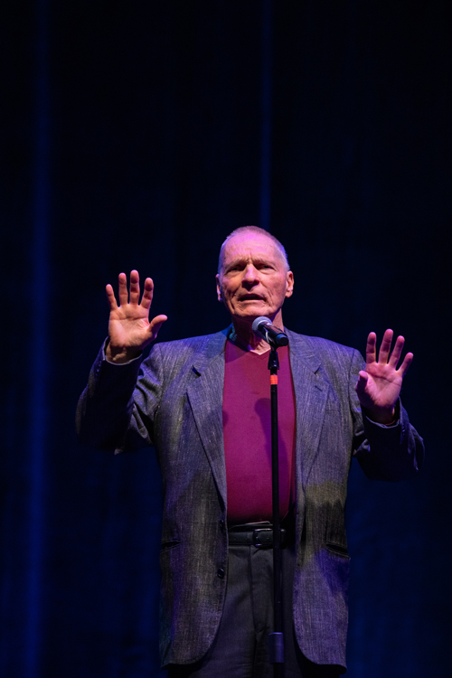 An expressive medium shot of an older man speaking or performing live on stage.Subject and PosePerson: An older man with light skin, short gray hair, and a clean-shaven face.Expression & Pose: He is looking slightly upward and forward with an intense, earnest expression, his mouth open as if speaking or mid-story.Gestures: He stands with both hands raised to chest-and-shoulder height, palms facing forward and fingers spread wide in a dramatic, open gesture to emphasize his words.Clothing and EquipmentAttire: He is dressed in a smart-casual style, wearing a dark gray textured blazer over a deep maroon V-neck shirt. His dark trousers are held with a black leather belt.Microphone: A microphone on a tall black stand is positioned directly in front of him, aligned with his chest. A small band of orange tape is visible near the top of the stand.Lighting and BackgroundLighting: The stage features high-contrast theatrical lighting. A warm spotlight illuminates the front of his face, shirt, and hands, while a cool blue and purple ambient wash highlights the sides of his blazer and the background.Background: The background is dark and minimalist, with subtle vertical textures from a stage curtain softly lit in deep blue tones.Photo credit: kmr studios