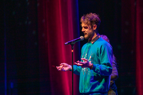 Subject: Scott Davis speaking intently into a microphone. Attire: A bright turquoise-blue crewneck sweatshirt worn over a dark collared shirt featuring a colorful patterned collar. Action & Expression: He is captured mid-speech with an expressive look on his face, holding both hands open in front of his chest in an engaging gesture. Setting & Lighting: Dramatic theatrical lighting highlights him against a deep red stage curtain on the left and cool dark shadows on the right. photo credit kmr studios