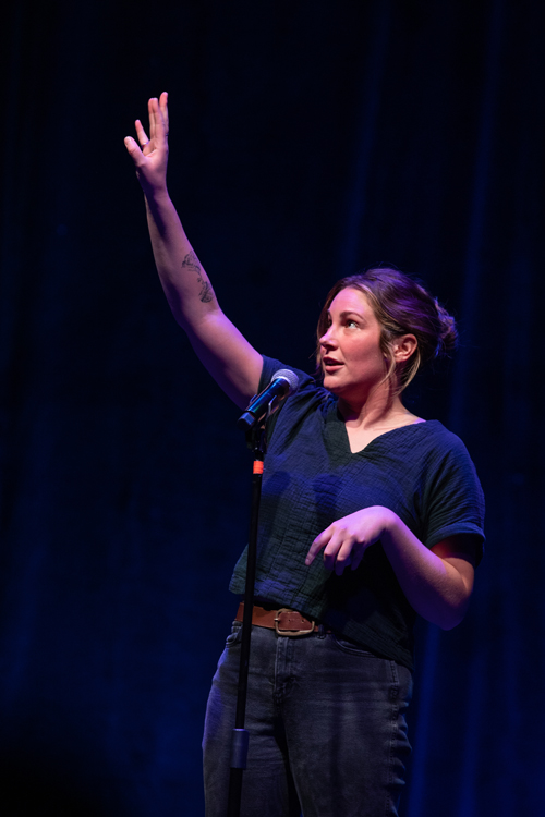 Subject: Ruthie Castle performing her story on stage. Attire: A dark slate-green short-sleeved top tucked into dark charcoal jeans with a brown leather belt. A flower tattoo is visible on her right forearm. Action & Expression: She reaches her right arm high above her head with an open palm while gesturing downwards with her left hand, speaking animatedly into the microphone. Setting & Lighting: Shot from a side angle, illuminated by a focused spotlight against a dark blue stage curtain. photo by kmr studios
