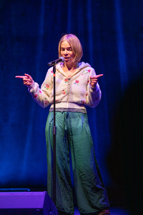 Subject: Emily Farrell sharing a lighthearted moment on stage. Attire: A cream-colored sherpa zip-up jacket decorated with embroidered red and blue flowers, paired with wide-leg teal drawstring trousers. Action & Expression: She stands in front of the microphone with a humorous, focused expression, pointing both index fingers outward in opposite directions. Setting & Lighting: Framed against a deep blue curtain backdrop, with a black floor stage monitor speaker visible in the bottom-left corner. photo by kmr studios