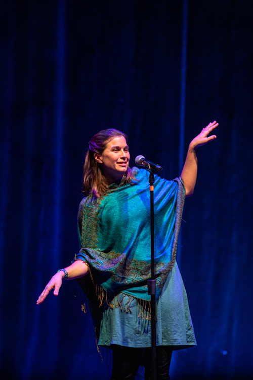 Subject: Shayla Paradeis gesturing dramatically during her performance. Attire: A teal tunic dress layered with a vibrant teal-and-gold paisley wrap/shawl featuring fringe trim, paired with dark leggings. Action & Expression: She smiles broadly while speaking into the microphone, extending her left hand high into the air and her right hand low in an open, welcoming gesture. Setting & Lighting: Illuminated under soft purplish-blue spotlights set against a textured dark curtain backdrop. photo by kmr studios