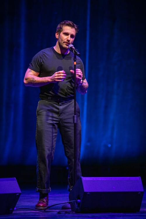 A full-length shot of a man speaking or performing live on a beautifully lit stage.Subject and PosePerson: A young man with short dark brown hair, a light mustache, and a trimmed beard. He has visible tattoos on both of his upper and lower arms.Expression & Pose: He is standing upright, facing slightly to his left, with a focused and expressive look as he speaks into the microphone.Gestures: His hands are raised near his chest, fingers slightly curved as if mid-sentence, bringing an active, storytelling energy to his stance.Clothing and EquipmentAttire: He is dressed in a casual, monochromatic style, wearing a fitted black crew-neck t-shirt tucked into dark gray or black trousers with a belt. The cuffs of his trousers are rolled up, revealing dark reddish-brown leather boots.Microphone & Audio: He stands directly behind a tall black microphone stand. Two large black stage monitor speakers sit on the floor in the foreground on either side of him.Lighting and BackgroundLighting: The stage is bathed in a dramatic, deep blue and purple glow. A warm spotlight highlights his face, arms, and upper body from the front, creating a strong contrast against the cooler ambient light.Background: Behind him is a large, softly textured stage curtain or backdrop illuminated by vertical streaks of blue light.Photo credit: kmr studios