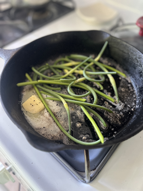 a high-angle, close-up shot of cooking in progress on a stovetop. Key Elements: The Pan: A well-seasoned black cast-iron skillet sits directly on a gas stove burner grate. The Ingredients: Inside the skillet, there is a handful of bright green garlic scapes (the curly, slender stalks harvested from garlic plants). They are curly, tangled, and tossed in the pan. The Cooking Liquid: A visible pool of melted, bubbling butter or oil occupies the bottom left side of the pan. A solid, rectangular pat of unmelted butter sits right in the middle of the pool, speckled with a bit of black pepper. The Background: The background is softly out of focus (shallow depth of field), showing parts of a white stovetop and kitchen counter area.