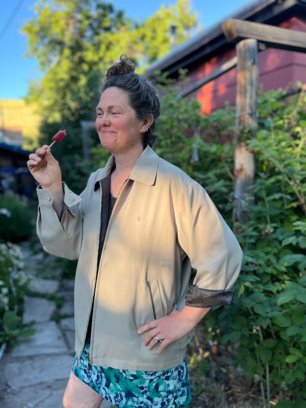 A medium shot captures a person, seemingly a woman, standing outdoors on a stone path, smiling and holding a small, red popsicle on a stick. Subject Details Appearance: The person has their dark hair styled up in a high bun. They are smiling warmly, looking slightly off-camera to the left. Clothing: They are wearing an oversized, light-beige zip-up jacket with a collar. The jacket has a small embroidered logo on the left chest and dark lining, which is visible where the left sleeve is cuffed. Underneath, they are wearing a dark top and green, white, and blue patterned floral shorts or a skirt. Pose: Their left hand rests on their hip, revealing a couple of rings on their fingers, while their right hand holds up the red popsicle. Background and Setting Environment: The setting appears to be a lush backyard or garden path. Green foliage, bushes, and trees fill the background. Structures: To the right, a wooden post and a portion of a red building or shed are visible. Lighting: The scene is illuminated by warm, late-afternoon sunlight casting gentle shadows, creating a relaxed, casual atmosphere.