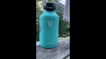 This animated GIF shows a teal or mint-green Hydro Flask insulated water bottle resting outdoors on a weathered wooden railing or deck. Object Details Brand/Logo: The white Hydro Flask stick-figure logo is prominently displayed on the front center, with the text "Hydro Flask" printed near the bottom base. Cap: It has a wide-mouth black cap with a built-in carrying strap. Condition: The bottle appears well-used, showing some scuffs, dark marks, and sticker residue on the front surface. A partial purple sticker can be seen on its left side. Background & Motion Motion: The GIF features a slight panning or rotating motion, giving a dynamic view of the water bottle. Setting: The background is softly blurred, showing green trees and natural outdoor lighting, suggesting it was taken in a backyard, park, or on a porch.