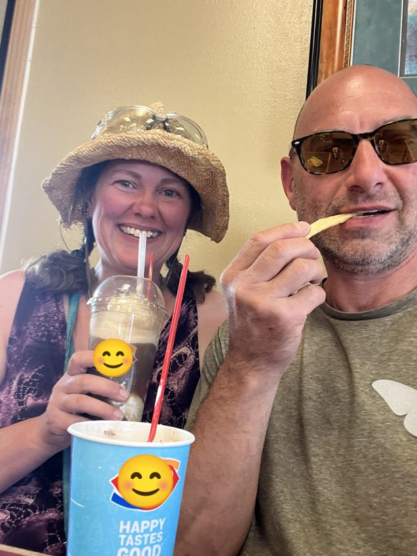 This selfie shows two people enjoying food and drinks indoors, likely at a Dairy Queen or similar casual restaurant. Key Details The Woman (Left): Wearing a wide-brimmed straw hat with sunglasses resting on top. Wearing a patterned, sleeveless dark purple top. smiling happily while sipping from a clear plastic cup filled with a blended drink or float (with a smiling emoji overlaid on the logo). The Man (Right): Bald, wearing dark tortoise-shell sunglasses and a olive-green t-shirt featuring a subtle graphic print on the chest. Has light facial stubble and is holding a single French fry up toward his mouth as if about to eat it. Foreground & Environment: In the lower center, there is a blue paper cup with the text "HAPPY TASTES GOOD" visible and a Dairy Queen style logo covered by a smiling emoji overlay. The background features plain, warm-toned walls and part of a framed picture frame hanging on the wall behind the man.