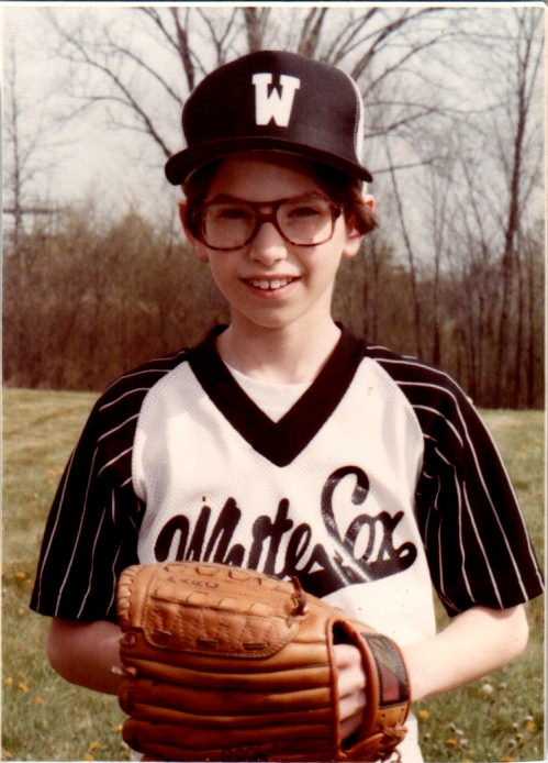 This is a vintage portrait of a young baseball player posed for an official team photo. Subject Details Attire: He is wearing a white baseball jersey with a black V-neck collar, black sleeves featuring thin white pinstripes, and a script logo across the chest that appears to read "White Sox." Cap: He wears a black baseball cap with a white letter "W" printed on the front. Accessories: He wears large, brown aviator-style prescription eyeglasses with dark frames, characteristic of late 1970s or early 1980s fashion. Equipment: He holds a brown leather baseball glove in front of him with both hands. The name "MARC" is hand-written in black ink on the thumb/pinky edge of the glove. Expression: He looks directly at the camera with a gentle, slight smile showing his upper teeth. Setting & Background Environment: He is outdoors in a grassy area scattered with dandelions. Background: In the background, there is a line of trees with bare or newly budding branches against a soft light sky, suggesting an early spring baseball season.