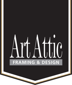 This image features the logo for Art Attic Framing & Design set inside a vertical banner shape. Banner Structure: The design is contained within a black shield- or banner-shaped frame featuring a pointed V-shaped bottom edge. It has a multi-layered border consisting of a thin outer gold line, a thick white inner border, and an accent line surrounding the dark background. Main Brand Name: Prominently centered in white is the text "Art Attic". It uses a stylized, tall condensed serif font with distinctive pointed crossbars and elegant, curly terminal accents (most notably on the lowercase "t"s and "c"). A solid white horizontal line runs directly beneath the name. Tagline: Directly under the underline, the phrase "FRAMING & DESIGN" appears in all-caps white lettering inside a solid light-grey rectangular bar.