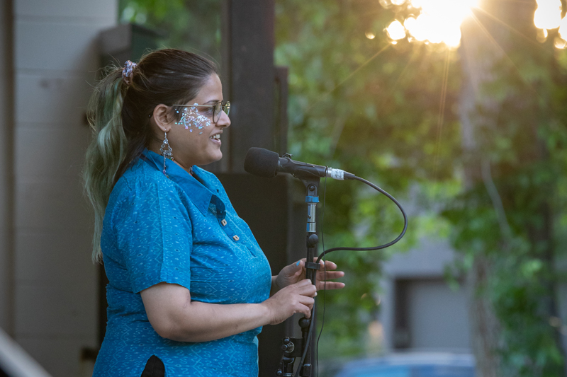 Subject: A profile/side-angle medium shot of a woman with her hair pulled back into a ponytail with green-tinted ends. She wears glasses, decorative glitter design on her cheekbone, and dangling earrings as she speaks into a microphone. Attire: She wears a vibrant turquoise patterned short-sleeve button collar shirt. Lighting & Setting: Set outdoors with warm, back-lit sunlight flares creating bokeh through green foliage in the background. Photo Credit: kmr studios