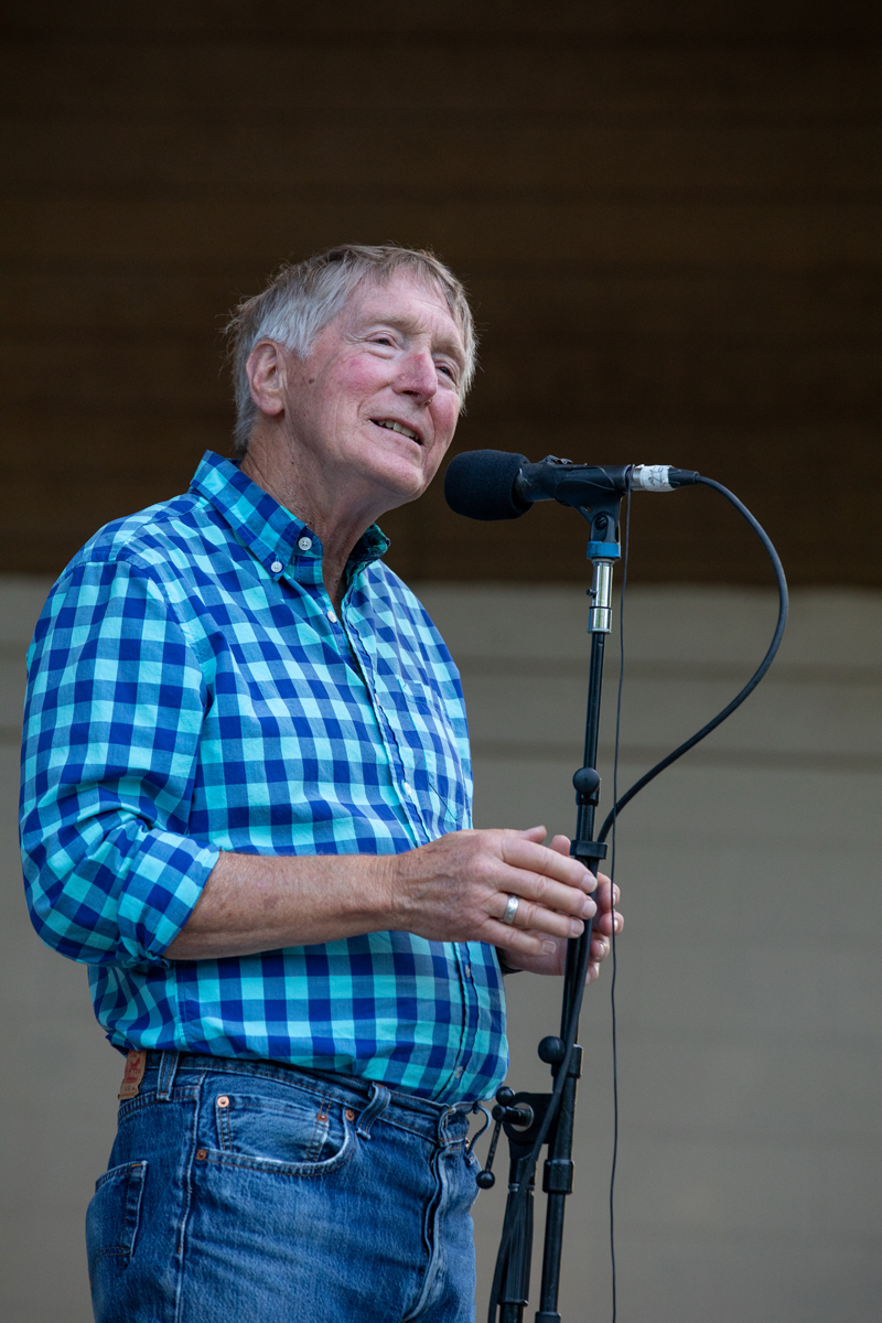 Subject: A medium close-up shot of an older man with short grey-brown hair smiling gently and looking upwards to the right while speaking into a microphone. His hands are lightly resting near the microphone stand. Attire: He wears a bright blue and teal checkered button-down shirt paired with blue jeans. Setting: On stage with a dark/neutral blurred background. Photo Credit: kmr studios