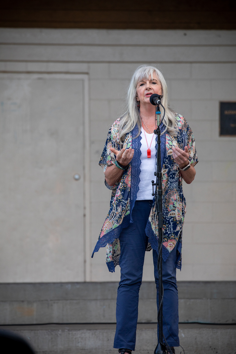 Subject: A full-length portrait of a woman with long, straight silver hair and bangs speaking into a microphone. She is gesturing outwards with open hands as she speaks. Attire & Accessories: She wears a white top under a flowy, navy blue floral-patterned kimono/cardigan with lace trim, dark blue trousers, a bright red whistle pendant necklace, and bracelets on her wrist. Setting: Standing on a stage in front of a neutral-colored brick/panel building backdrop. Photo Credit: kmr studios