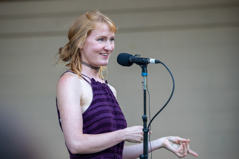 Subject: A medium close-up shot of a strawberry-blonde/red-haired woman smiling warmly while speaking at a microphone. Her hair is styled in a loose side updo, and she gestures naturally with one hand open toward the audience. Attire & Accessories: She wears a dark purple textured halter top/dress, a black lace choker necklace, and has a small geometric tattoo visible on her inner palm. Setting: Indoors or on a covered stage in front of a light-colored neutral wall. Photo Credit: kmr studios
